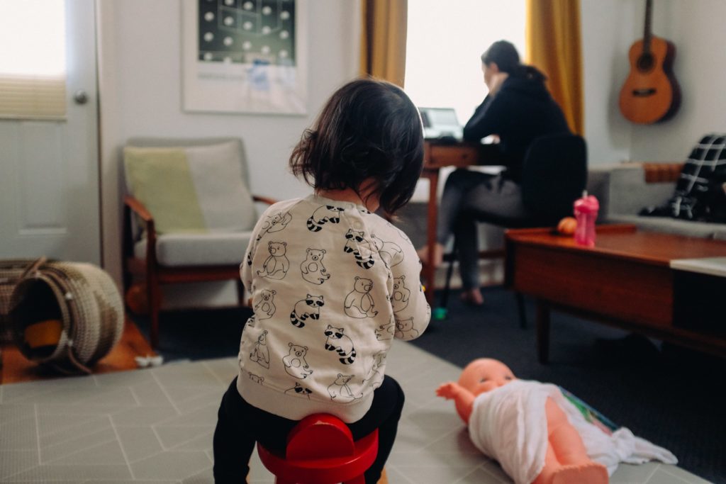 A person works in the background on a laptop at a desk while a child plays in the foreground